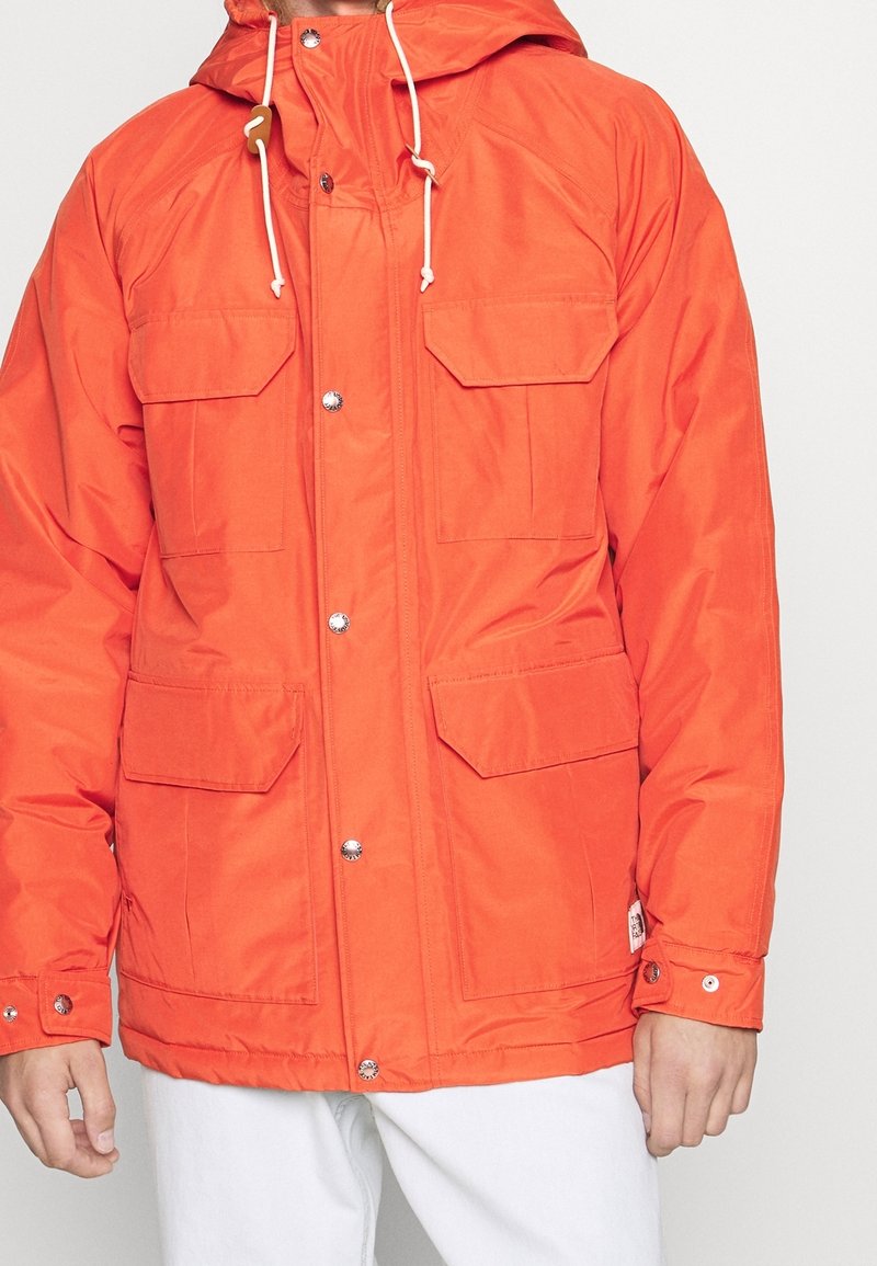 Homme portant une veste à capuche orange vif avec des boutons-pression à l'avant et quatre poches à rabat, associée à un pantalon blanc.