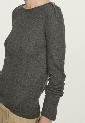 Mørkegrå langærmet sweater med knapdetalje på skulderen, båret med lyse bukser, vist fra nakke til hofter.