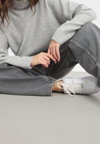 Grå stickad tröja med hög hals, löst sittande grå denimbyxor och vita sneakers med texturerad yta och snördetaljer.