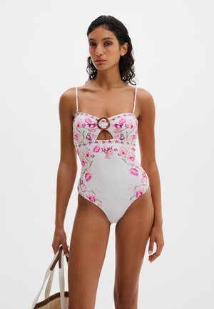 Nainen pukeutuneena valkoiseen kukkakuosiseen uimapukuun, jossa on kilpikonnakuviollinen rengasdetaili, pitäen beigeä olkalaukkua, jossa on valkoiset hihnat, yhtenäistä taustaa vasten.