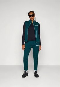 Turquoise sportief vest en broekset gemaakt van lichtgewicht stof, met een vest met rits, lange mouwen en zwarte sneakers.