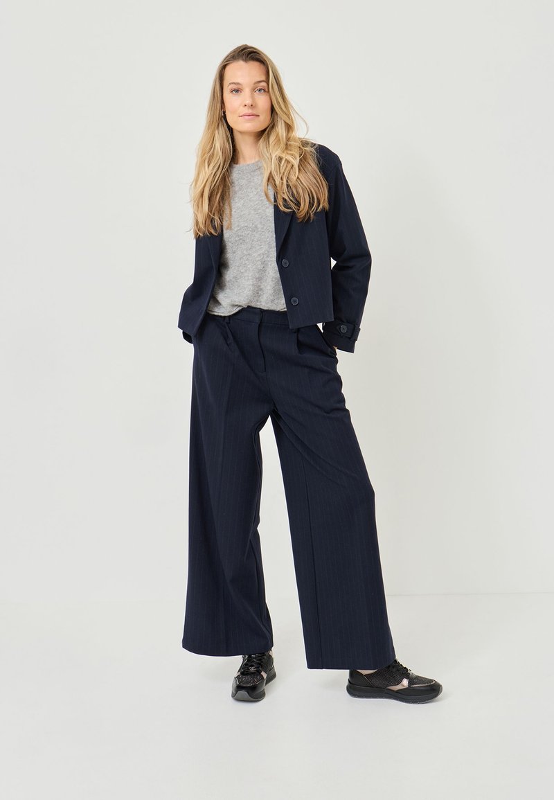 Veste de costume rayée bleu marine sur un pull gris, associée à un pantalon large et des baskets noires. Le tissu semble lisse avec des boutons standards.