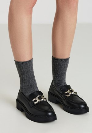 Mocassins en cuir noir avec détail en chaîne argentée, semelle épaisse et finition lisse, assortis à des chaussettes côtelées grises.