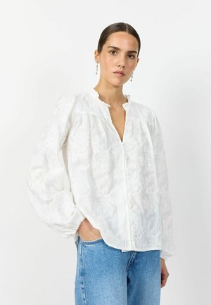 Femeie purtând o bluză albă brodată cu flori, cu mâneci bufante și blugi albastru deschis, stând în fața unui fundal alb simplu.