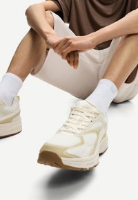Svetlo béžové športové topánky s panelmi z sieťoviny a gumovou podrážkou, spárované s bielymi rebrovanými ponožkami, nosené so svetlo sivými šortkami.