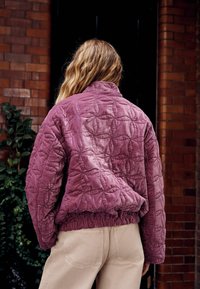 Steppjacke aus weichem, staubigem Violettstoff mit hohem Kragen, elastischem Saum und diamantförmigen Details. Kombiniert mit hellen Hosen.