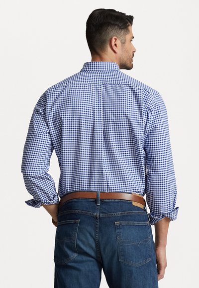 Blå och vit rutmönstrad skjorta med uppvikta ärmar, kombinerad med mörka jeans och ett brunt bälte, sedd bakifrån.