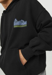 Felpa nera con tasca a marsupio, polsini a coste e una stampa grafica in blu e verde che rappresenta edifici e il testo "PARKS&Leisure®."