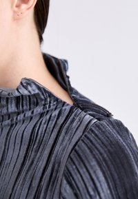Tamsiai pilka klostuota palaidinė su aukštu kaklu, pasižyminti raukšlėta tekstūra ir mygtukų užsegimu nugaroje. Lygi, blizgi medžiaga su sluoksniuotais raukšlėmis.
