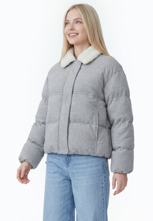 Jeune femme portant une doudoune gris clair avec un col en polaire blanche et un jean bleu clair, souriante et regardant légèrement sur le côté.