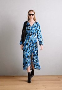 Dlouhé květinové šaty v modrých tónech s překládaným designem a dlouhými rukávy. Spárovány s černými kotníkovými botami a texturovanou černou kabelkou.
