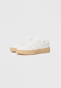 Hvite sneakers laget av et glatt, læraktig materiale og en teksturert beige gummisåle. Har snøring og subtil branding på siden.