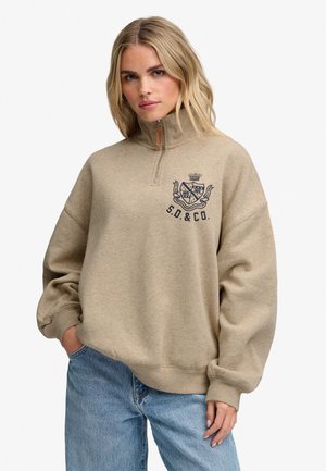 Vrouw met blond haar die een beige sweater met kwart rits en S.D.&Co.-logo en blauwe jeans draagt, staat met één hand in de zak.