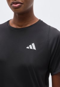 Κοντινό πλάνο ενός ατόμου που φοράει μαύρο αθλητικό μπλουζάκι με λευκό τρίγωνο λογότυπο με τρεις ρίγες στο στήθος.