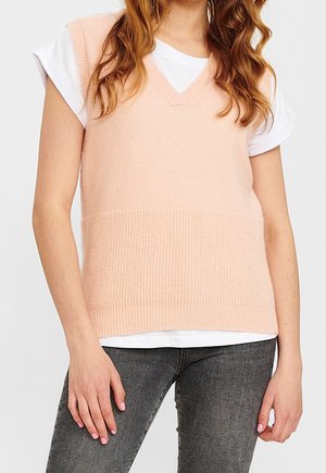 Kobieta w bezrękawniku z jasnoróżnego dzianego materiału na białej koszulce i ciemnoszarych dżinsach, stojąca z luźno opuszczonymi rękami.