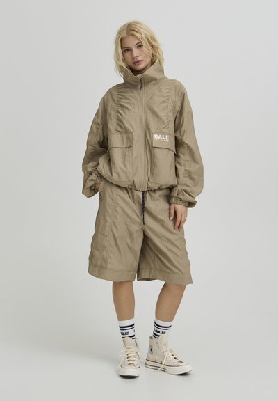 Veste beige courte avec fermeture éclair, deux poches avant, et poignets élastiques, associée à un short assorti jusqu'aux genoux. Baskets blanches avec accents bleus.