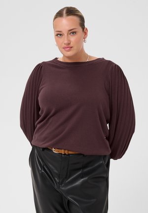 Femme portant un pull violet foncé à manches plissées, un pantalon en cuir noir et une ceinture marron, debout les mains dans le dos.
