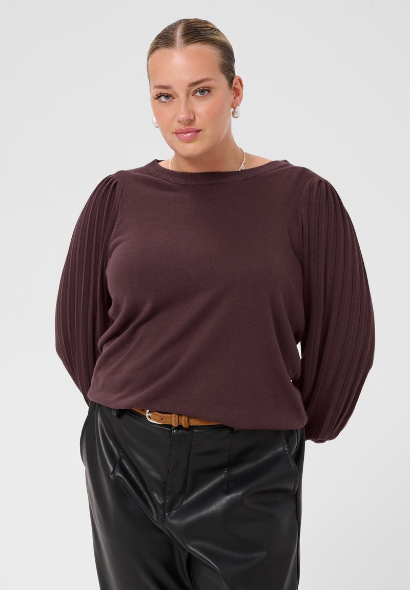 Femme portant un pull violet foncé à manches plissées, un pantalon en cuir noir et une ceinture marron, debout les mains dans le dos.