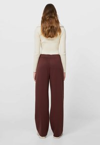 Bruine wijde broek van een gladde stof met een elastische tailleband, gecombineerd met een aansluitende crèmekleurige geribbelde trui. Gezien vanaf de achterkant.