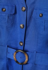 Gros plan sur un vêtement en tissu bleu avec quatre boutons marron, deux poches poitrine et une ceinture assortie avec une boucle ronde en écaille de tortue.