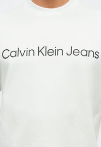 Λευκό μπλουζάκι με την επιγραφή "Calvin Klein Jeans" σε μαύρο πάνω στο στήθος, φορώντας το άτομο που φαίνεται από το λαιμό έως το μέσο του κορμού.