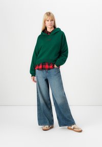 Haut à capuche vert avec un logo sur le devant, superposé à une chemise écossaise rouge et noire. Un jean bleu à jambe large et des chaussures claires complètent la tenue.