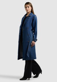 Naine, kes kannab pikkade varrukatega sinist teksatrenchi mustade pükste ja kontsakingadega, seisab lihtsa halli tausta ees.