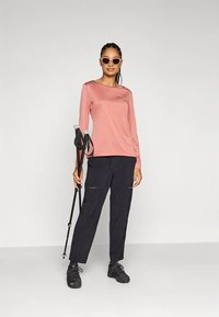 Žena u sunčanim naočalama, ružičastoj dugoj majici, crnim hlačama i planinarskim cipelama drži crne planinarske štapove ispred jednobojne pozadine.