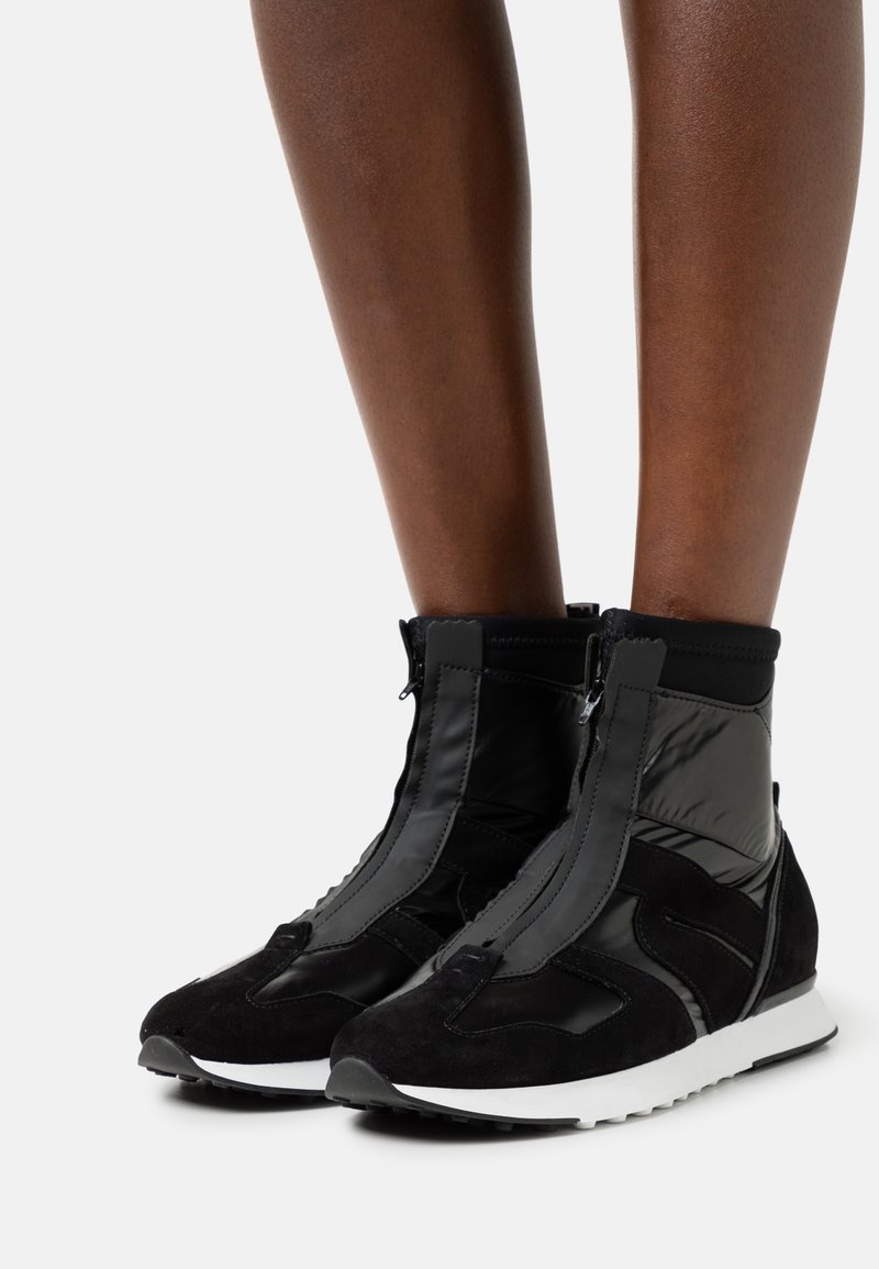 Jambes portant des baskets montantes noires avec fermeture éclair frontale, design en daim et panneaux brillants, et semelles en caoutchouc blanches sur un fond clair.