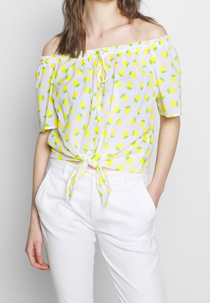 Frau trägt eine weiße schulterfreie Bluse mit gelbem Zitronenmuster, die in der Taille gebunden ist, kombiniert mit einer weißen Hose, eine Hand in der Tasche.