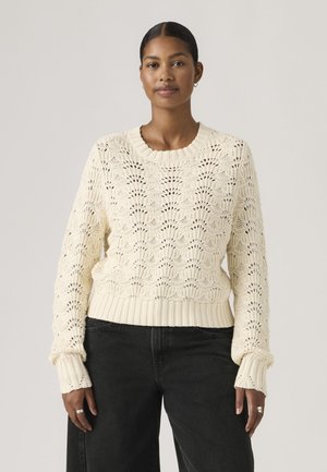 Kvinde med mørkt hår trukket tilbage iført cremefarvet åbenstrikket sweater og sorte bukser, stående mod en ensfarvet lys baggrund.