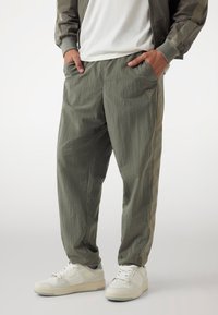 Lichte olijfgroene broek met een gestructureerde afwerking, voorzien van zijzakken en een logo-opdruk op het linkerpand, gecombineerd met witte sneakers.