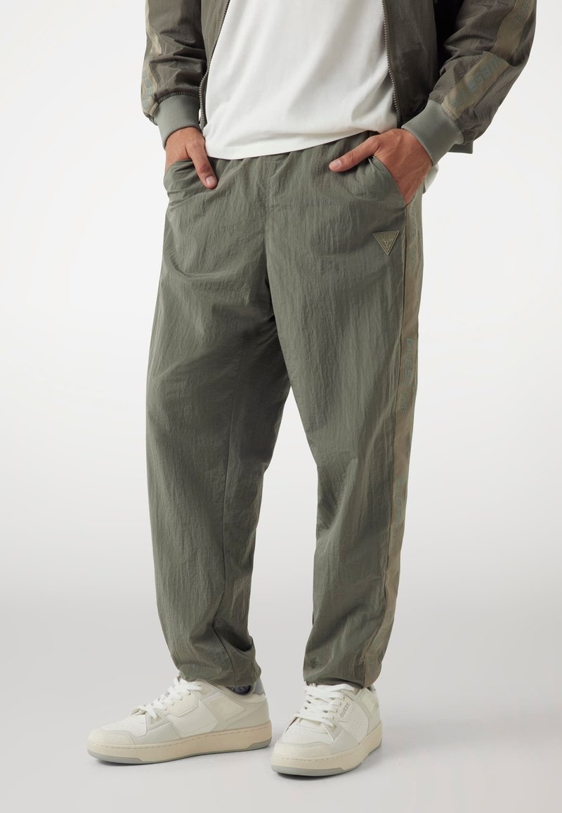 Lichte olijfgroene broek met een gestructureerde afwerking, voorzien van zijzakken en een logo-opdruk op het linkerpand, gecombineerd met witte sneakers.