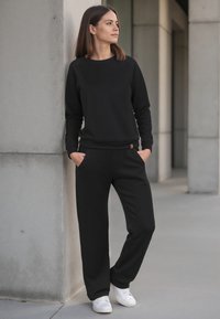 Sudadera negra y pantalones deportivos a juego, hechos de un tejido suave. Corte relajado con puños de canalé. Manos en los bolsillos. Zapatillas blancas como calzado.