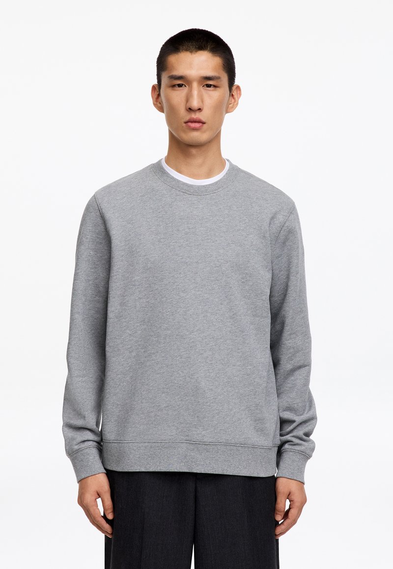 Homme aux cheveux courts et noirs portant un sweat-shirt gris uni à col rond et un pantalon noir, debout devant un fond blanc.