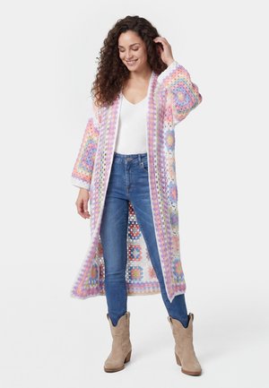 Frau lächelt und berührt ihr Haar, trägt blaue Jeans, weißes Top, beige Stiefel und eine lange pastellfarbene mehrfarbige gehäkelte Strickjacke.