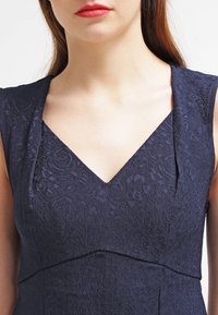 Middendonk blauw mouwloos jurkje met een gestructureerd bloemenpatroon, een V-halslijn en een gestructureerd lijfje, voorzien van subtiele plooidetails rond de hals.