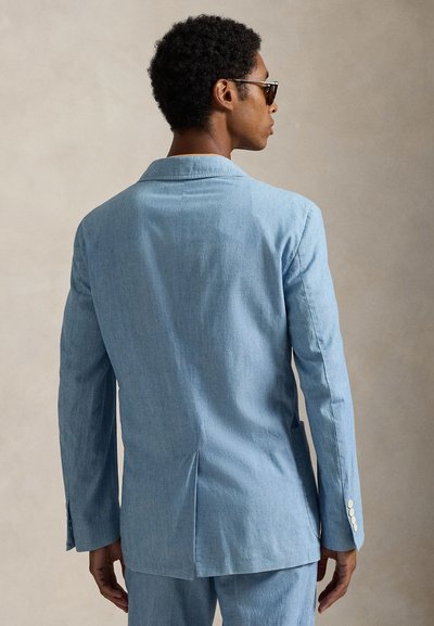 Ανοιχτό μπλε denim σακάκι με μακριά μανίκια, δύο πλαϊνές τσέπες και λευκές λεπτομέρειες με κουμπιά, με σκίσιμο στην πλάτη για ευκολία κινήσεων.