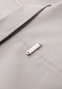 Gros plan sur un tissu beige avec des coutures détaillées et une petite étiquette métallique rectangulaire argentée gravée du mot "QUINTESSENCE."
