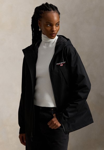 Svart vanntett jakke med glidelås foran, justerbar hette og logo på brystet. Brukt over en hvit høyhalset genser. Enkelt, funksjonelt design.