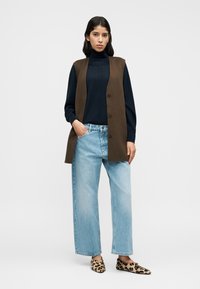 Καφέ γιλέκο χωρίς μανίκια πάνω από μπλε μπλούζα με ψηλό λαιμό, σε συνδυασμό με ανοιχτό μπλε παντελόνι με φαρδιά μπατζάκια και παντόφλες με λεοπάρ σχέδιο.