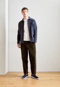 Marineblaues Hemdjacke mit Vordertasche, weißes T-Shirt, olivgrüne Cordhose und marineblaue Slipper mit weißen Sohlen.