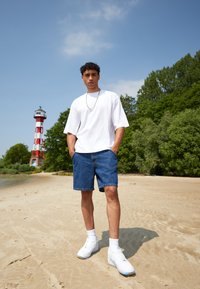 Tricou alb supradimensionat, pantaloni scurți din denim și adidași albi. Fundal de plajă cu nisip, un far cu dungi roșii și albe și vegetație.