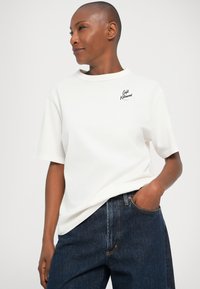 T-shirt bianco in cotone con maniche corte, colletto rotondo e logo nero sul petto, abbinata a jeans in denim blu scuro, mani in tasca.