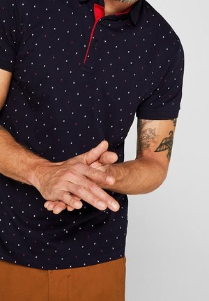 Marineblaues Poloshirt mit einem Mikroquadratmuster in Weiß und Rot, roten Kragenakzenten und kurzen Ärmeln. Der Stoff wirkt glatt mit einer leichten Textur.