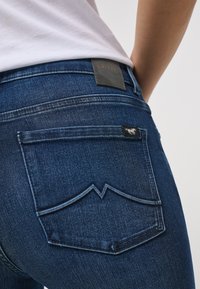 Tmavě modré džíny z denimu s výrazným designem zadní kapsy, který má špičatý šev. Štítek je z černé kůže s embosovaným textem.