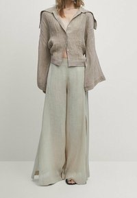 Beige cropped overhemd met knopen, lange, brede mouwen en strikjes op de schouders; gecombineerd met lichtgroene, wijde broek met zijsplitten.