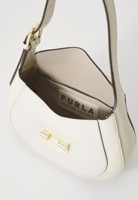 Kremowa skórzana torebka Furla z złotym zapięciem, otwarta u góry z widoczną etykietą wewnętrzną "Furla Genuine Leather."