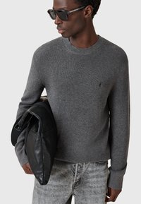 Szary sweter z prążkowanego dzianiny z okrągłym dekoltem, połączony z ciemnymi dżinsami i trzymający czarną skórzaną torbę. Model nosi ciemne okulary przeciwsłoneczne.