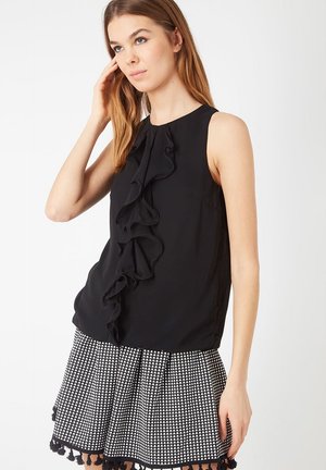 Joven mujer con cabello largo que lleva una blusa sin mangas negra con volantes y una falda a rayas en blanco y negro con flecos.